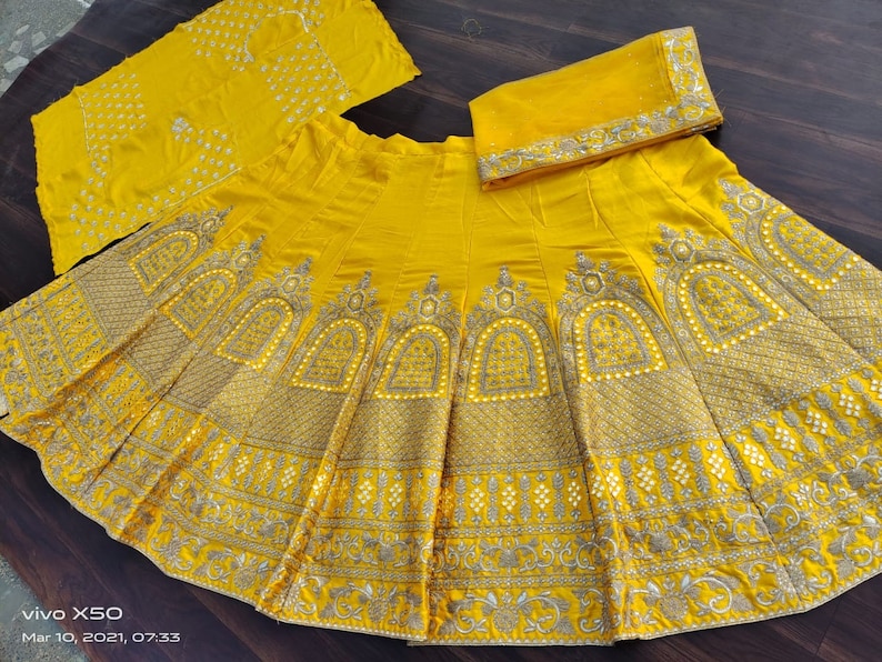 May include: A yellow skirt with intricate gold embroidery and embellishments. The skirt is made of a soft, flowing fabric and has a flared silhouette. The skirt is paired with a matching yellow dupatta, which is a long scarf that is often worn with Indian clothing.