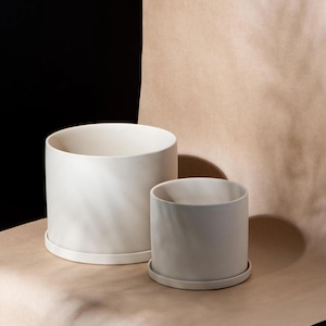 May include: Two white ceramic plant pots of different sizes, each with a matching saucer. The pots are cylindrical with a smooth, matte finish. The background is a mix of brown paper and black.