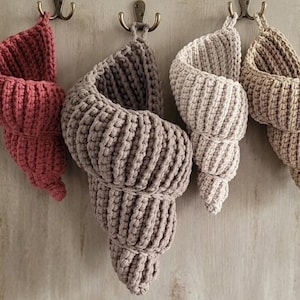 Könnte beinhalten: Vier handgefertigte, gehäkelte Wandkörbe in Muschelform in Rot-, Grau-, Beige- und Beigetönen. Jeder Korb wird an einem Metallhaken aufgehängt. Die Körbe sind aus dickem Garn gefertigt und haben eine gerippte Textur.