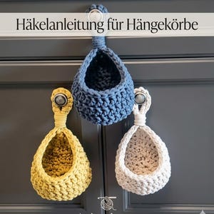 Può includere: Tre cestini sospesi all'uncinetto fatti a mano in giallo, blu e bianco sporco. Ogni cestino è sospeso da un pomello di metallo. Il testo "Häkelanleitung für Hängekörbe" è nella parte superiore dell'immagine.