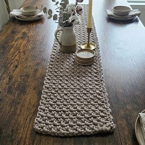 Könnte beinhalten: Ein rechteckiger, beigefarbener, gehäkelter Tischläufer mit strukturiertem Maschenmuster, mittig auf einem dunkelbraunen Holztisch platziert. Der Läufer hat ein kleines lederartiges Etikett und ist mit einem Keramikkrug, getrockneten Baumwollzweigen, einer Kerze und Untersetzern dekoriert.