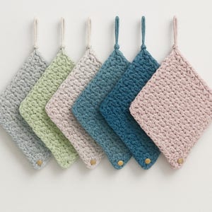 Könnte beinhalten: Satz von sechs gehäkelten Topflappen in verschiedenen Blautönen, Grüntönen und Rosa. Jeder Topflappen hat einen kleinen, runden goldenen Knopf.