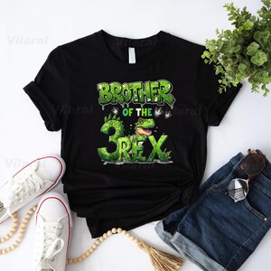 May include: Black t-shirt with a green graphic that reads "BROTHER OF THE 3 REX." The design features a cartoon dinosaur and the number 3. The shirt is displayed with white sneakers, denim shorts, and sunglasses.