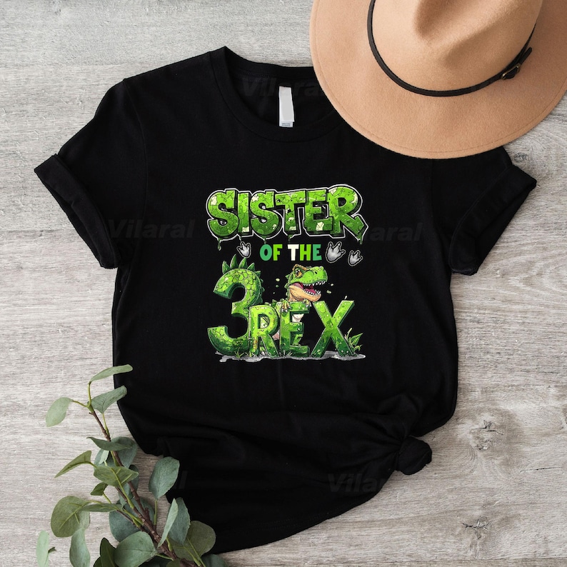May include: Black t-shirt with a green graphic that reads "SISTER OF THE 3 REX". The design features a dinosaur and the number 3 in a green, textured font. A tan hat is visible in the background.