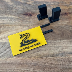 May include: A yellow rectangular sign with a black snake illustration and the text "no step on snek." Two black stands and two black rods are also in the image. The objects are on a wooden surface.