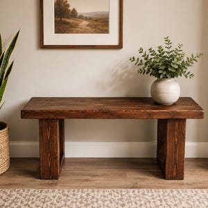 May include: A rectangular wooden bench with a dark brown stained finish and a visible wood grain pattern. The bench features a thick, flat seat supported by two sturdy, wide block-style legs on each end, creating a simple and functional design.