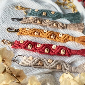 Könnte beinhalten: Fünf bunte Makramee-Schlüsselanhänger mit Holzperlen, die die Namen Hazel, Juliet, Coco buchstabieren. Jeder Schlüsselanhänger hat einen goldenen Schlüsselring aus Metall.