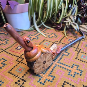 May include: A wooden smoking pipe with a carved snake design. The pipe is brown and black and has a long stem. The pipe is sitting on a colorful rug.