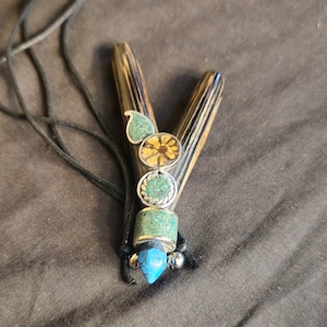 May include: A black and brown wooden pendant with turquoise and gold accents. The pendant is attached to a black cord.