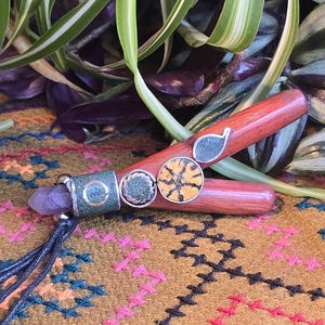 May include: A wooden pendant with a purple amethyst stone, green and gold accents, and a black cord. The pendant is on a brown and colorful patterned fabric.