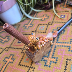 May include: A wooden pipe with a carved frog design. The frog is brown and green with blue eyes. The pipe is resting on a brown and gold patterned surface.