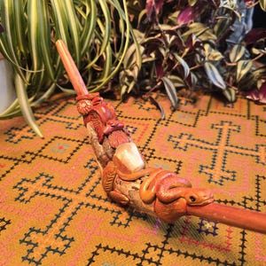 May include: A hand-carved wooden pipe with a snake design. The pipe is painted in shades of orange and brown. The snake is wrapped around the pipe and has a detailed head.