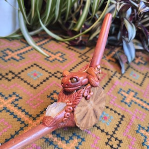 May include: A hand-crafted wooden pipe with a ceramic frog figurine on top. The pipe is decorated with a clear crystal and intricate details.