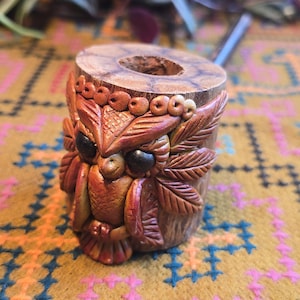 May include: A small wooden pipe carved in the shape of an owl with brown, orange, and black details. The owl has large eyes and a detailed feather pattern.