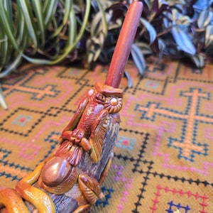 May include: A handcrafted wooden pipe with intricate carvings and a rich, warm color palette. The pipe features a detailed design with a snake, leaves, and other decorative elements. The stem is a deep reddish-brown, and the overall aesthetic is earthy and artistic.