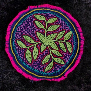 May include: A circular embroidered design with a green leafy pattern in the center, surrounded by a pink, blue, and yellow border.