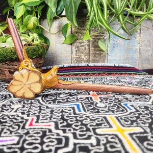 May include: A wooden smoking pipe with a carved bird head and a flower-shaped bowl. The pipe is resting on a colorful patterned fabric.