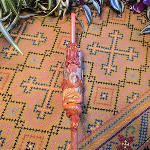 May include: A wooden pipe with a carved snake design. The snake is red and brown and has a detailed face. The pipe is on a patterned fabric background.