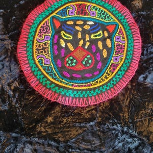 Op de afbeelding: Een ronde, geborduurde patch met een gestileerd jaguar gezicht. Het ontwerp bevat een zwarte achtergrond met kleurrijke details in rood, groen, geel, paars en blauw. De patch heeft een roze, gefranjerde rand.