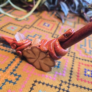 May include: A wooden pipe with a carved snake design. The snake is wrapped around the pipe and has a red and brown color scheme. The pipe is resting on a patterned fabric.