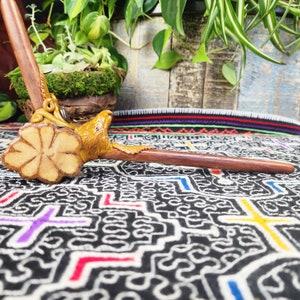 May include: A handcrafted wooden pipe with a carved eagle head and floral design. The pipe is made of dark brown wood with a light brown eagle head and floral accents. The pipe is displayed on a patterned fabric with plants in the background.