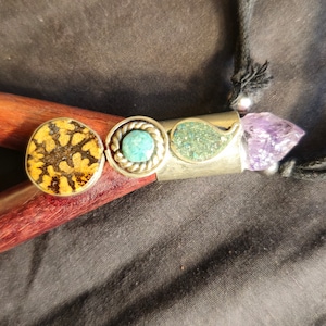 May include: A handcrafted necklace featuring a silver pendant with three decorative elements: a round, patterned stone, a turquoise stone, and a leaf-shaped green stone. The necklace also includes a purple crystal and a black cord.