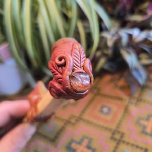 May include: A close-up of a handmade clay pen with a decorative top featuring a swirl design and a clear quartz crystal. The pen is painted in shades of brown and red.