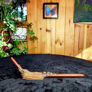 May include: A wooden smoking pipe with a brown and red finish. The pipe is shaped like a tree branch with a carved owl on the side. The pipe is sitting on a black surface.