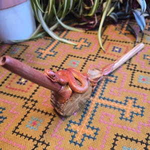 May include: A wooden pipe with a carved snake design. The pipe is brown and has a red snake wrapped around it. The snake's head is at the top of the pipe and its tail is at the bottom. The pipe is sitting on a patterned rug.