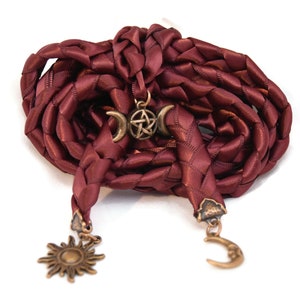 May include: A burgundy braided cord with three metal charms: a crescent moon, a pentagram, and a sun. The cord is braided from a satin-like material.