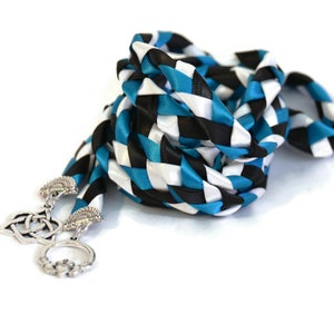 May include: A braided cord with a black, white, and teal checkered pattern. The cord has two silver Celtic knot charms attached to the ends.