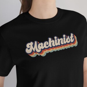 Puede incluir: Una camiseta negra con un gráfico de estilo retro que dice "Machinista" en colores arcoíris.
