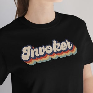 Op de afbeelding: Een zwart T-shirt met een retro-stijl grafische afbeelding met de tekst "Invoker" in regenboogkleuren.