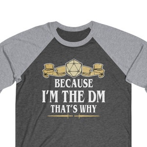 May include: A dark heather grey and light grey baseball tee shirt with a white print that reads "Because I'm the DM That's Why". The print also features a gold and white illustration of a 20-sided die.