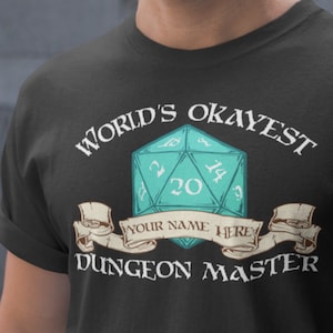 May include: Black t-shirt with a white graphic of a 20-sided die and the text "World's Okayest Dungeon Master". The die is teal and has white numbers. The text "Your Name Here" is on a banner below the die.