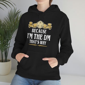 Op de afbeelding: Zwarte hoodie met een witte afbeelding met de tekst "Because I'm the DM That's Why" met een gouden dobbelsteen en een scrollontwerp.