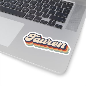 May include: A retro-style sticker with the name "Tauren" in a colorful, wavy font. The sticker has a white background and is printed with a multi-colored design.
