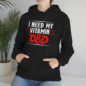 Könnte beinhalten: Schwarzer Hoodie mit einem weißen Aufdruck, der "Ich brauche mein Vitamin D&D" mit einem roten Drachen- und Würfeldesign sagt.