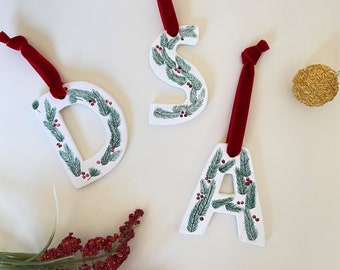 May include: Three white ceramic ornaments shaped like the letters D, S, and A. Each letter is decorated with green pine branches and red berries. The ornaments are hung with red velvet ribbon.