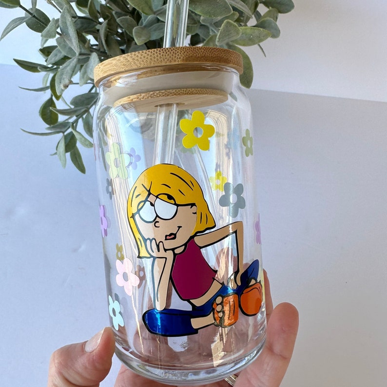 May include: A clear glass jar with a bamboo lid and a straw. The jar has a cartoon illustration of a girl with blonde hair wearing a red shirt and blue pants. The jar is decorated with colorful flowers.