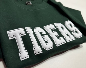 May include: A dark forest green crewneck sweatshirt featuring the word TIGERS printed in large, bold, white block letters with a thin white outline across the chest. The garment has a ribbed collar and long sleeves, made of a smooth fabric.