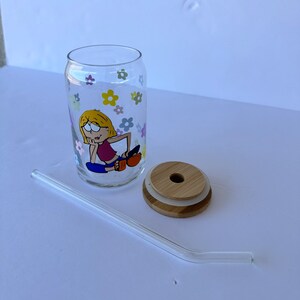 May include: A glass drinking cup with a cartoon illustration of a girl sitting with flowers around her. The cup has a bamboo lid with a silicone ring and a clear glass straw.