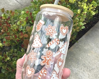 May include: A clear glass tumbler with a bamboo lid and a glass straw, featuring a repeating pattern of white and orange holiday-themed icons including gingerbread people, candy canes, snowflakes, mittens, hearts, and Christmas trees.