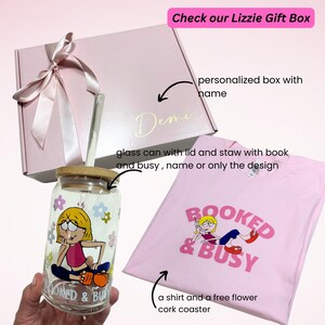 May include: A pink gift box with a white ribbon and the name "Demi" written on it. The box contains a glass can with a lid and straw, a pink t-shirt with the text "BOOKED & BUSY" and a cartoon illustration of a woman on a phone, and a cork coaster.