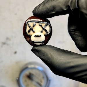 May include: A brown and white glass bead with a monkey face design. The bead features black accents for the eyes and mouth. The bead is held between two gloved fingers.