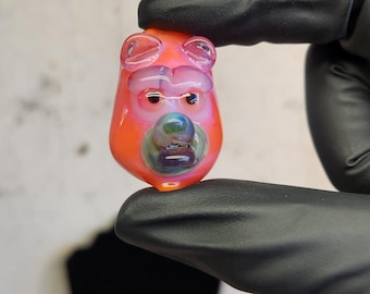 May include: A handmade glass pendant in the shape of a bear's head. The pendant is primarily orange with pink and blue accents. The bear has a grumpy expression. The pendant is held by a person wearing black gloves.