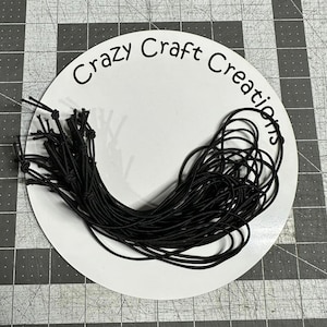 May include: A close-up of a white circle with the text "Crazy Craft Creations" printed in black. A large bundle of black elastic string is on top of the circle.