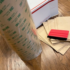 K&ouml;nnte beinhalten: Eine Rolle braunes 3M Klebeband mit gr&uuml;ner Beschriftung, ein roter Rakel und mehrere Bl&auml;tter braunes Papier.