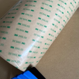 K&ouml;nnte beinhalten: Eine Rolle braunes Klebeband mit gr&uuml;nem Text, auf dem wiederholt "3M 300LSE" steht. Im Vordergrund befindet sich ein blauer Kunststoffrakel mit einem schwarzen Gummiband.