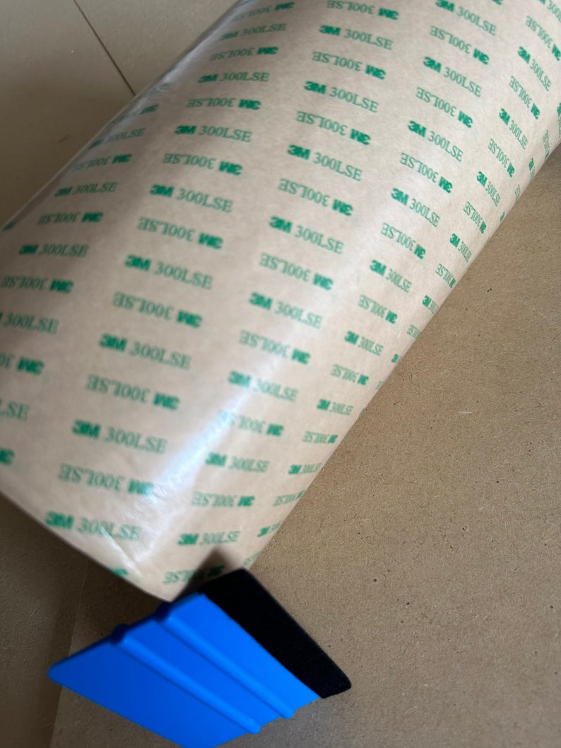 K&ouml;nnte beinhalten: Eine Rolle braunes Klebeband mit gr&uuml;nem Text, auf dem wiederholt "3M 300LSE" steht. Im Vordergrund befindet sich ein blauer Kunststoffrakel mit einem schwarzen Gummiband.